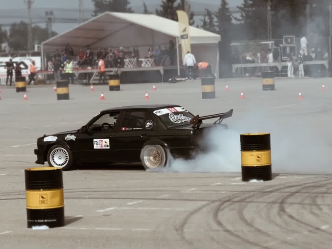 A black coupé sliding sideways past a row of oil drums, tyre smoke trailing behind it.
