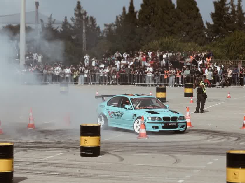 A pale blue coupé with a tall rear wing laying smoke in front of a packed crowd.