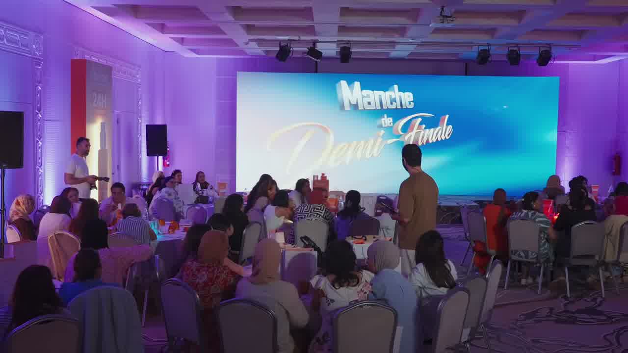 An audience seated at round tables facing a lit stage screen, the room washed in pink and purple light.