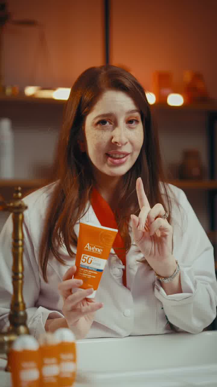 A woman in a white coat holding up an orange Avène sunscreen tube behind a counter, shelves of product behind her.