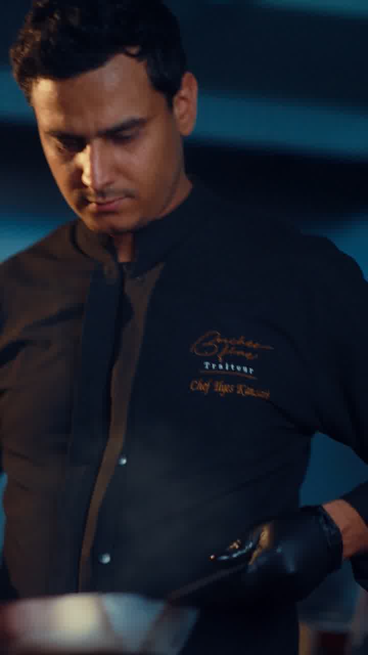A chef in an embroidered black chef’s jacket working over a pan in a dark kitchen.