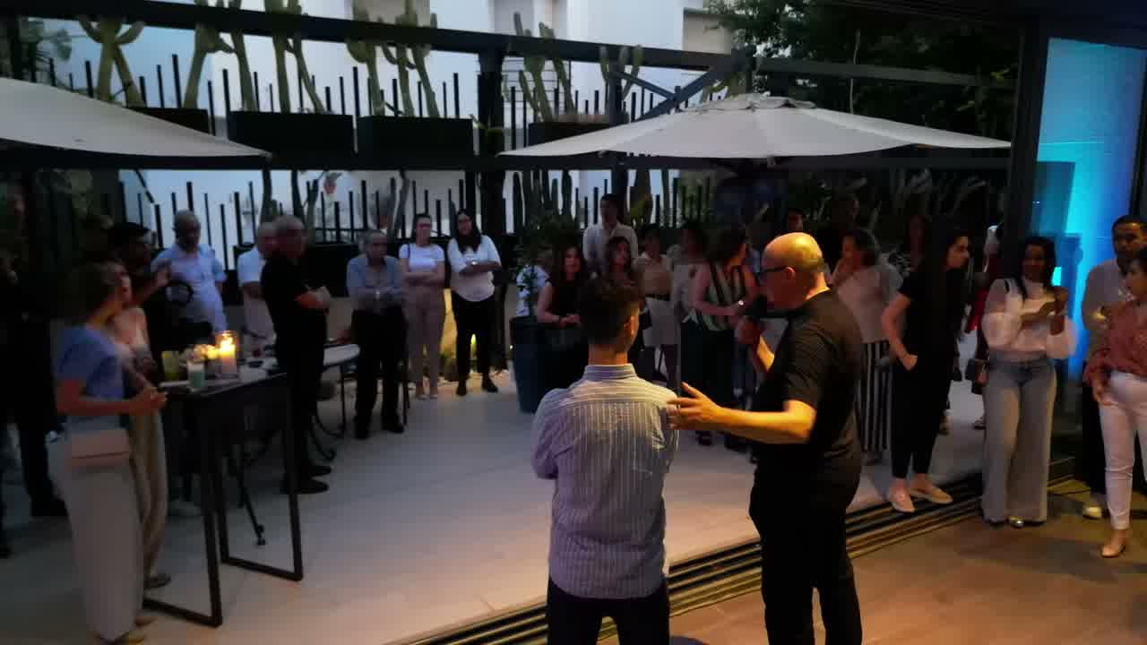 Guests standing and talking on a planted terrace at dusk, under parasols and warm lighting.