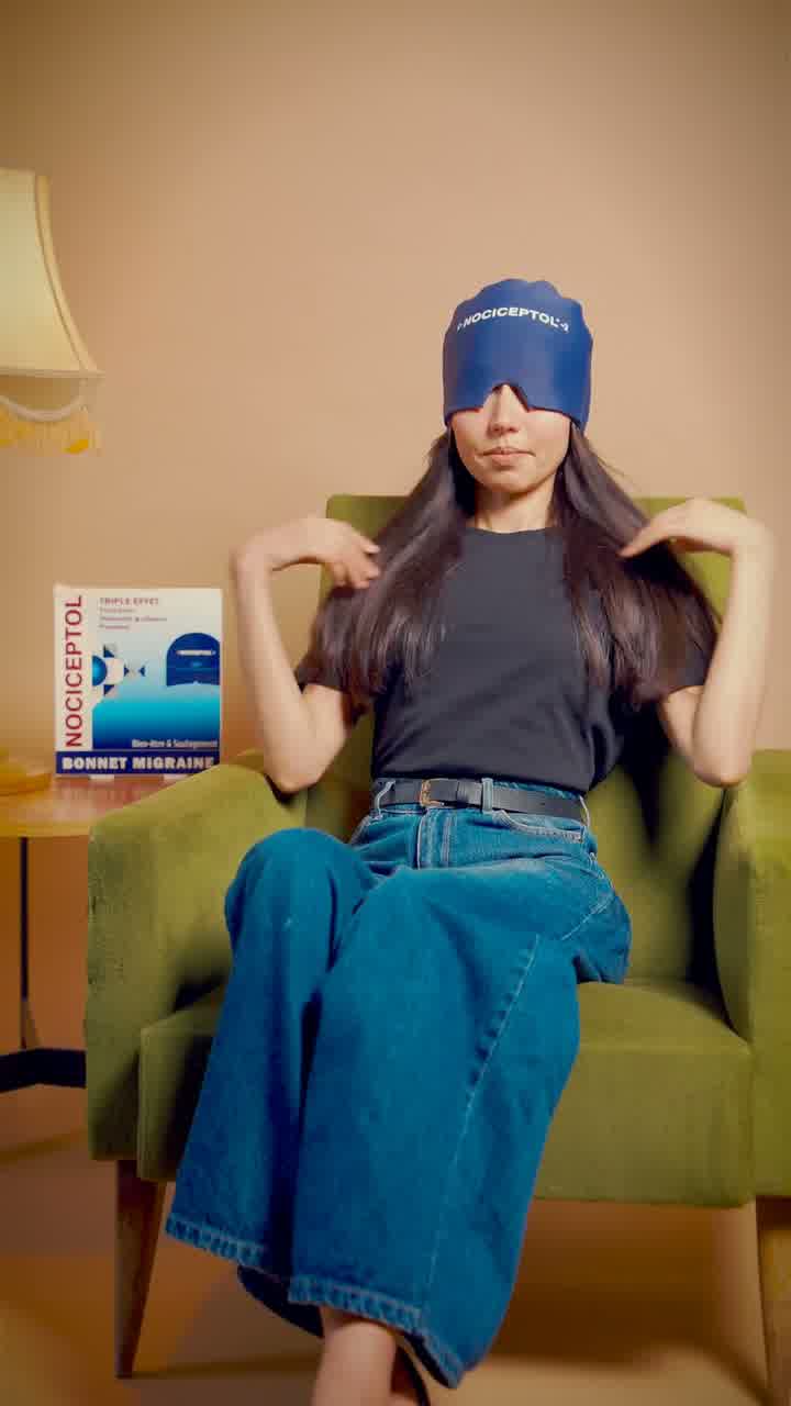 A woman in a green armchair wearing a navy Nociceptol eye mask, the product box resting on the chair arm.