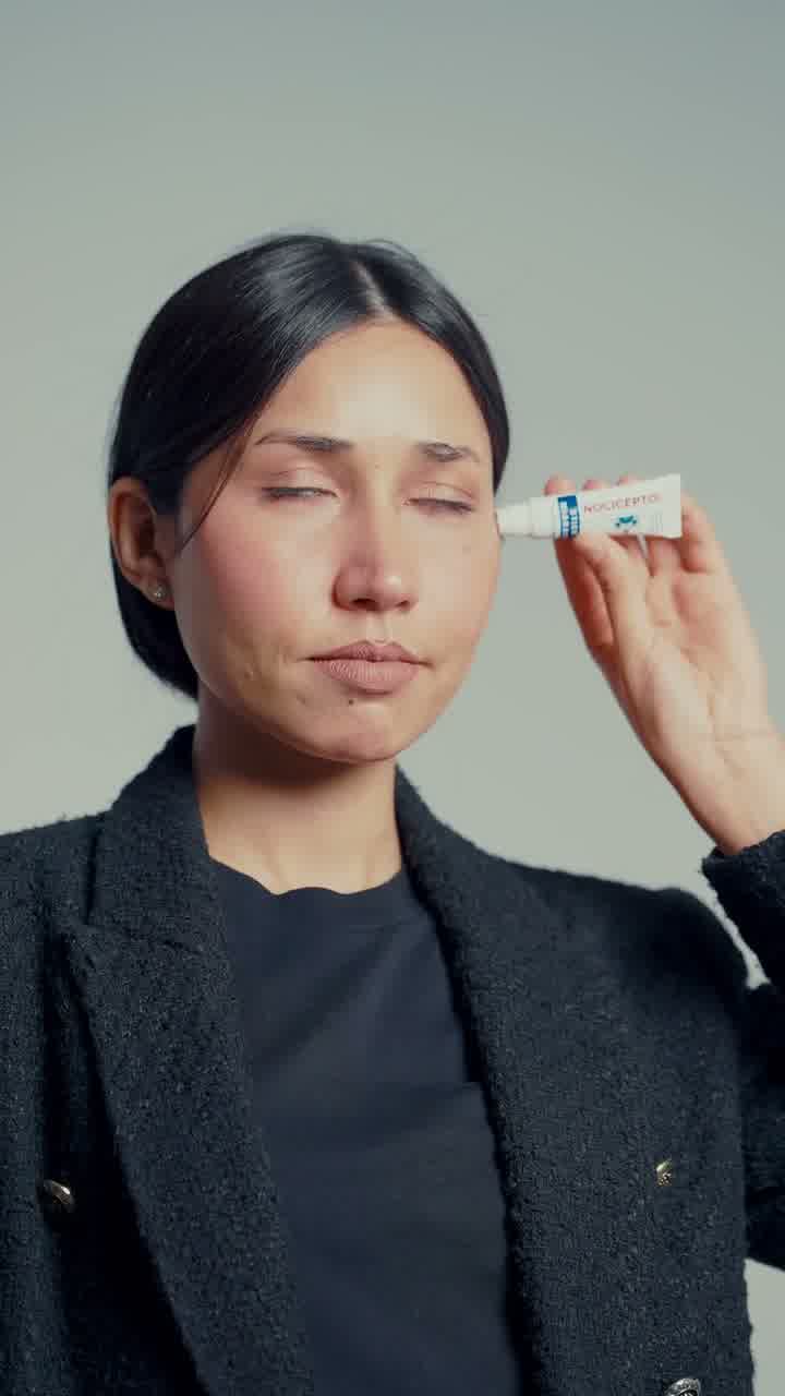 A woman with her eyes closed holding a small tube to her temple, against a dark background.