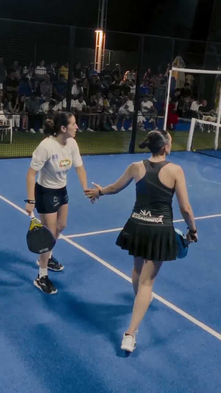 Two players on a floodlit blue padel court, with spectators behind the glass wall.