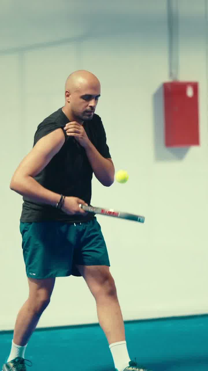 A player mid-swing on an indoor padel court, the ball in the air beside him.