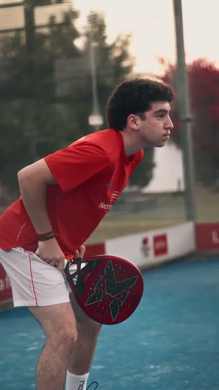 A young player in a red shirt crouched low with a padel racket, waiting to receive.