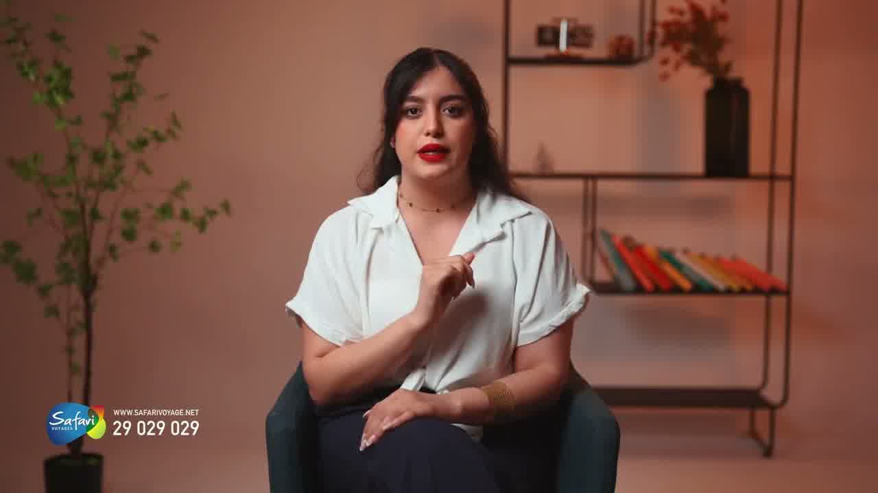 A presenter in a white shirt seated against a pink wall with shelving and a plant, gesturing to camera.