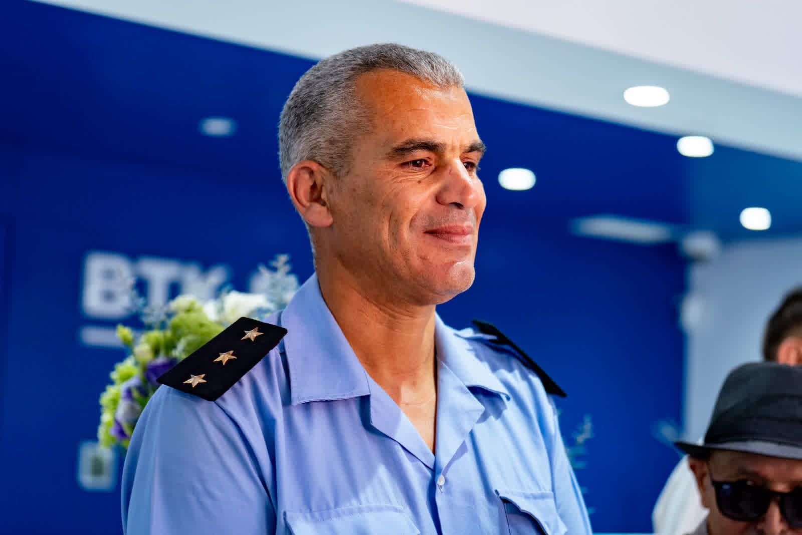 A man in a light blue uniform shirt speaking, a lit blue wall behind him.