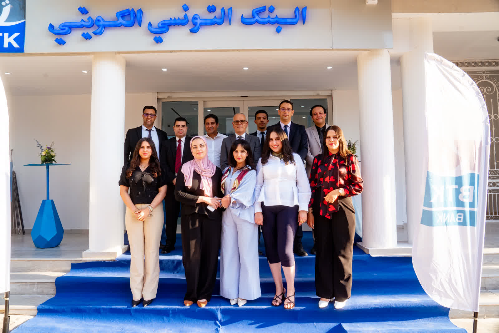 A group standing on a blue carpet at the entrance to a building, under an Arabic sign.