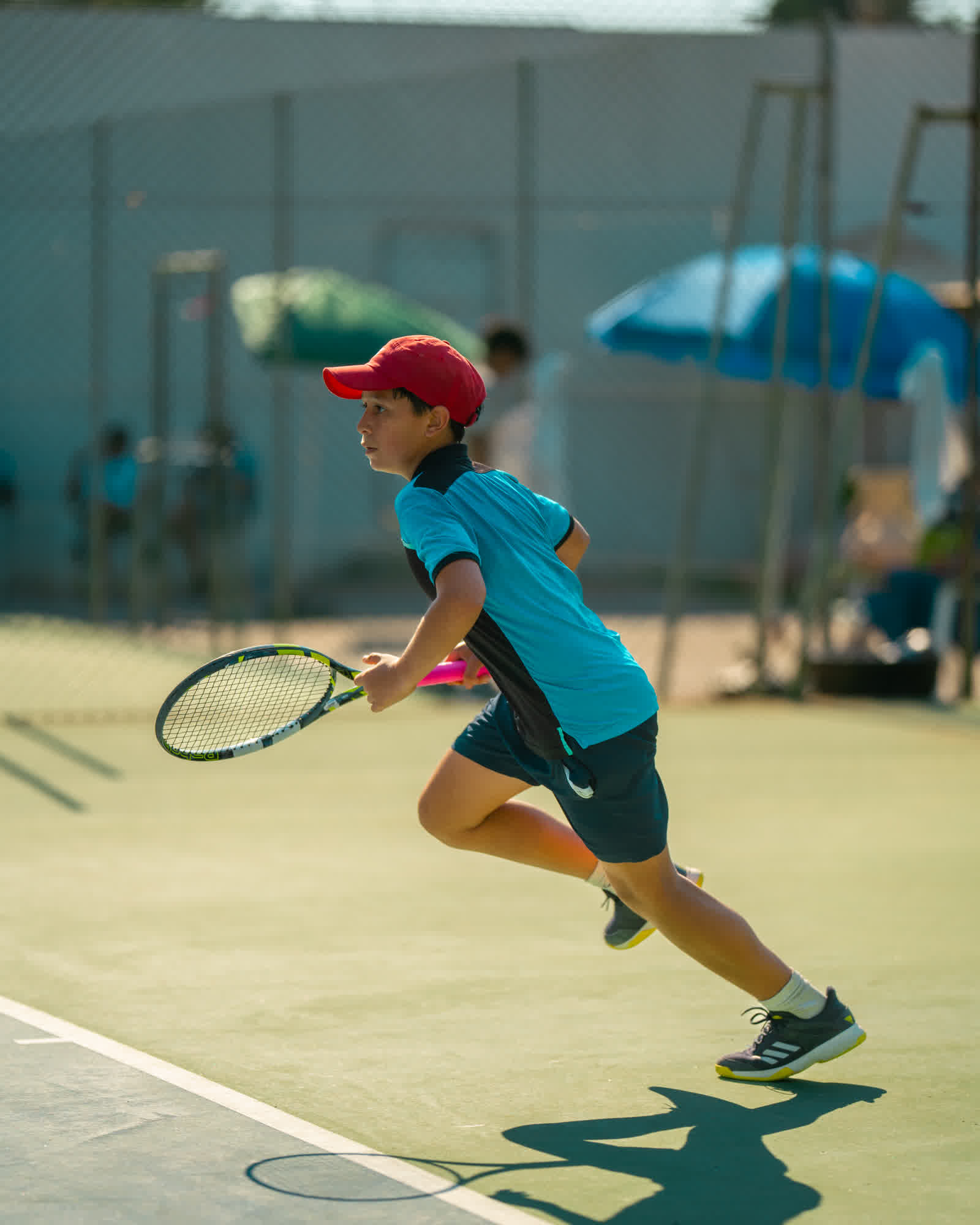 A young player in a red cap running across a clay court, racket in hand.