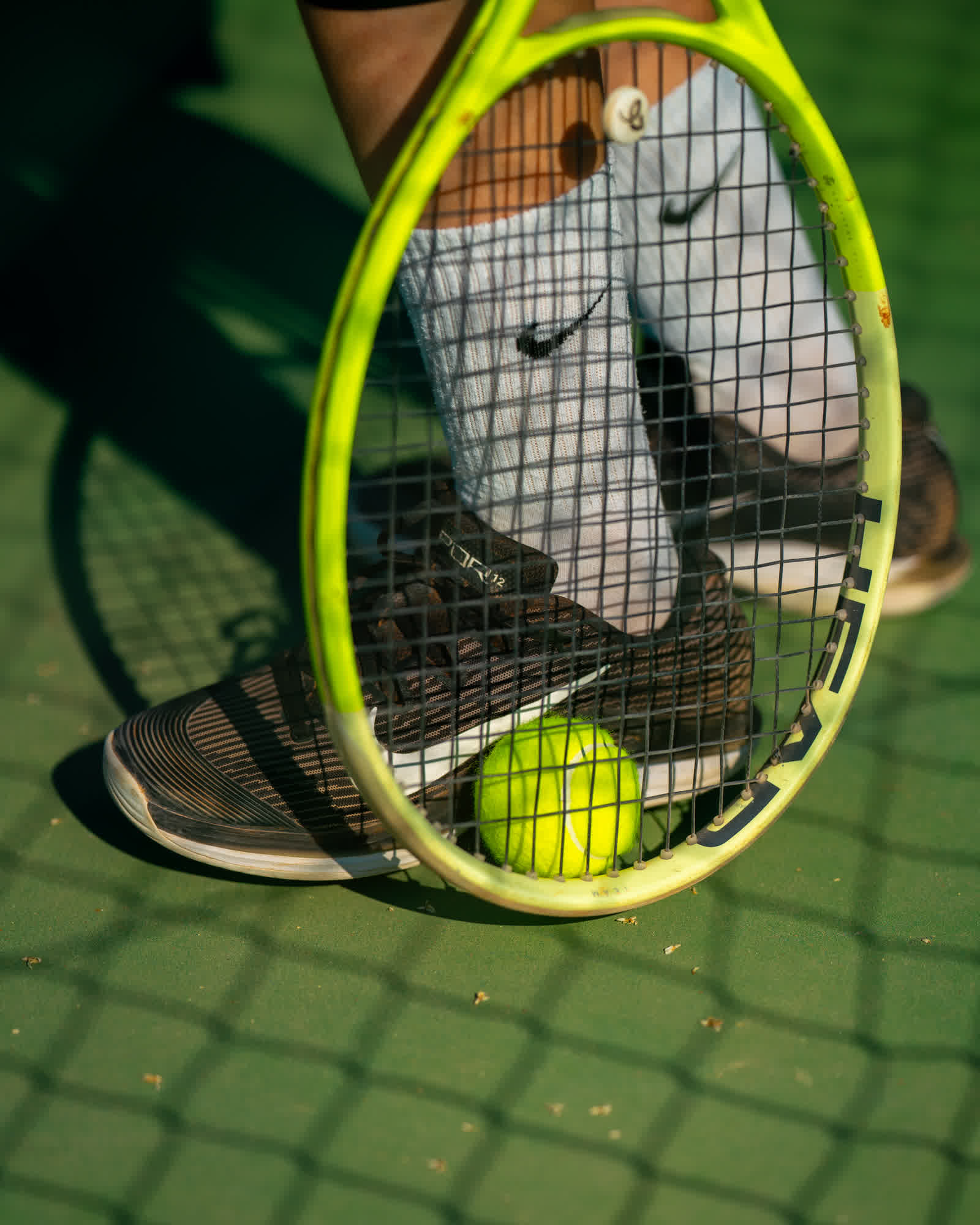 A racket and ball resting on the ground beside a player’s feet, seen through the strings.