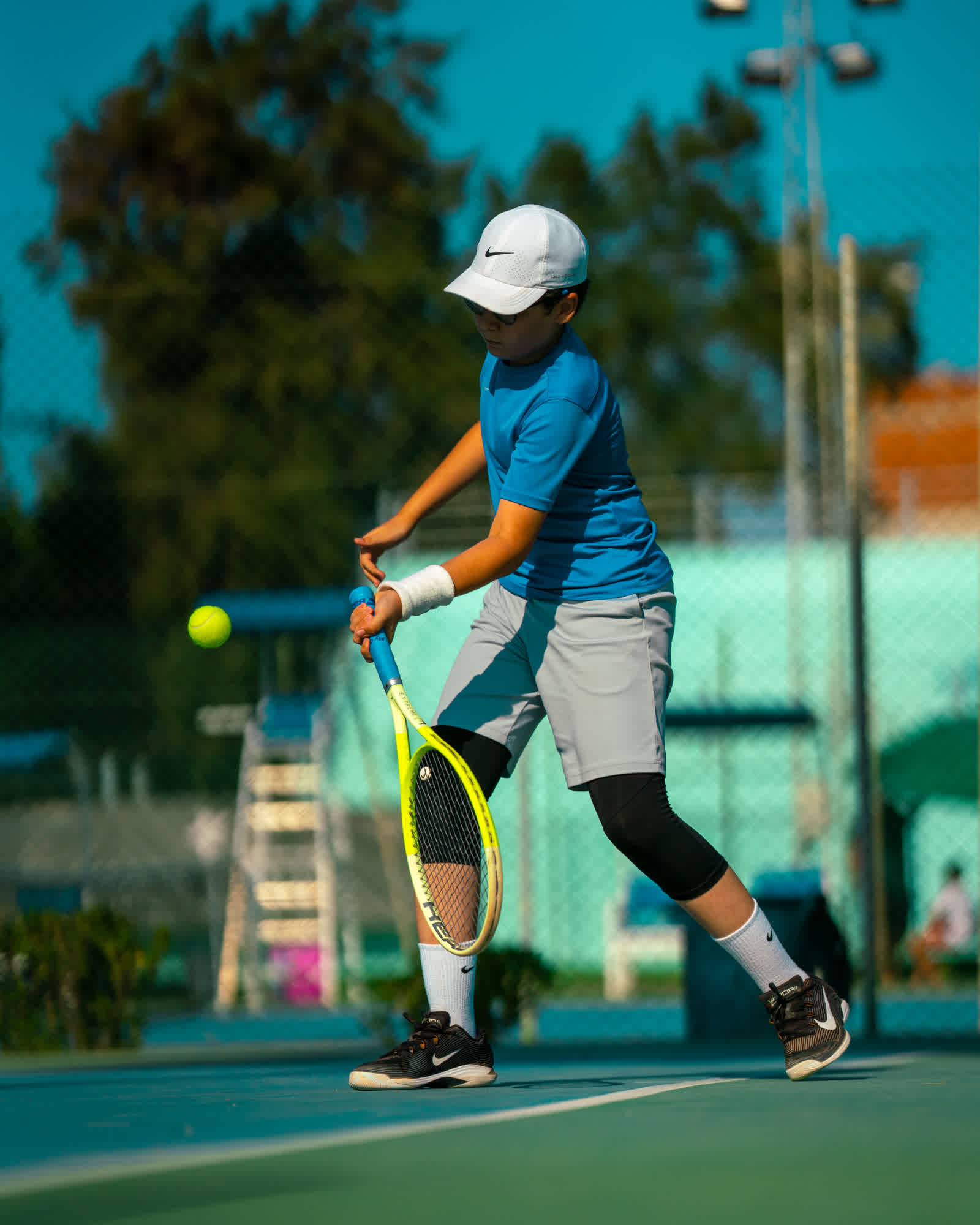 A young player in a white cap and blue shirt preparing to serve, ball in hand.