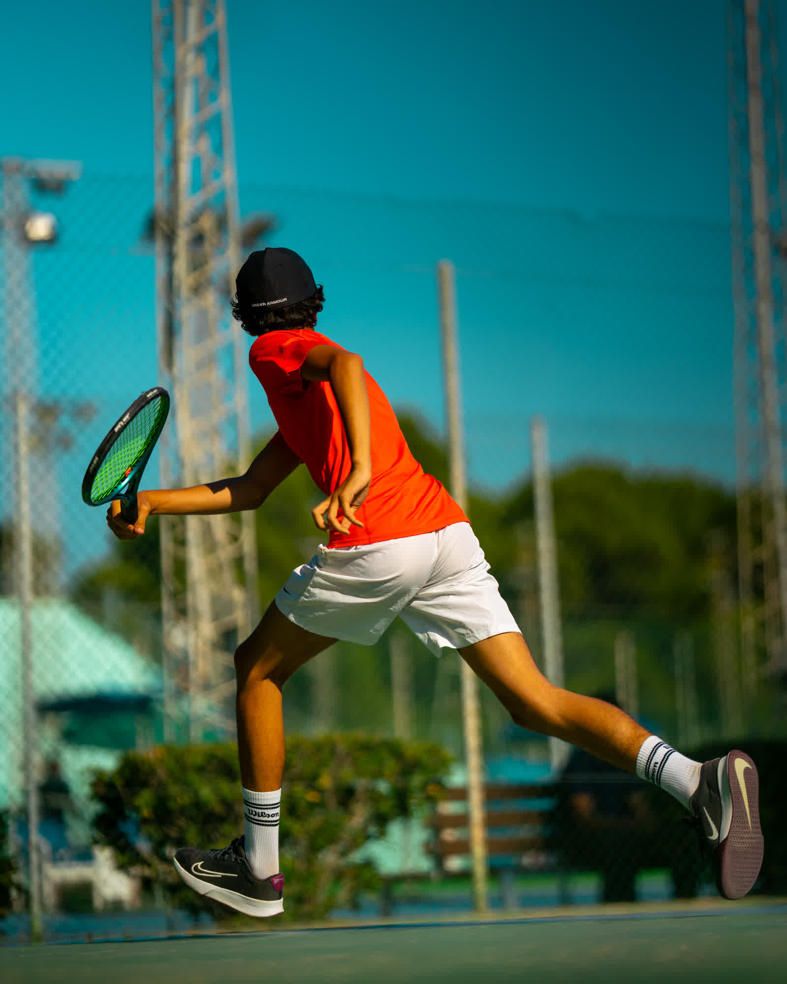 A young player in an orange shirt stretching into a forehand, both feet off the ground.