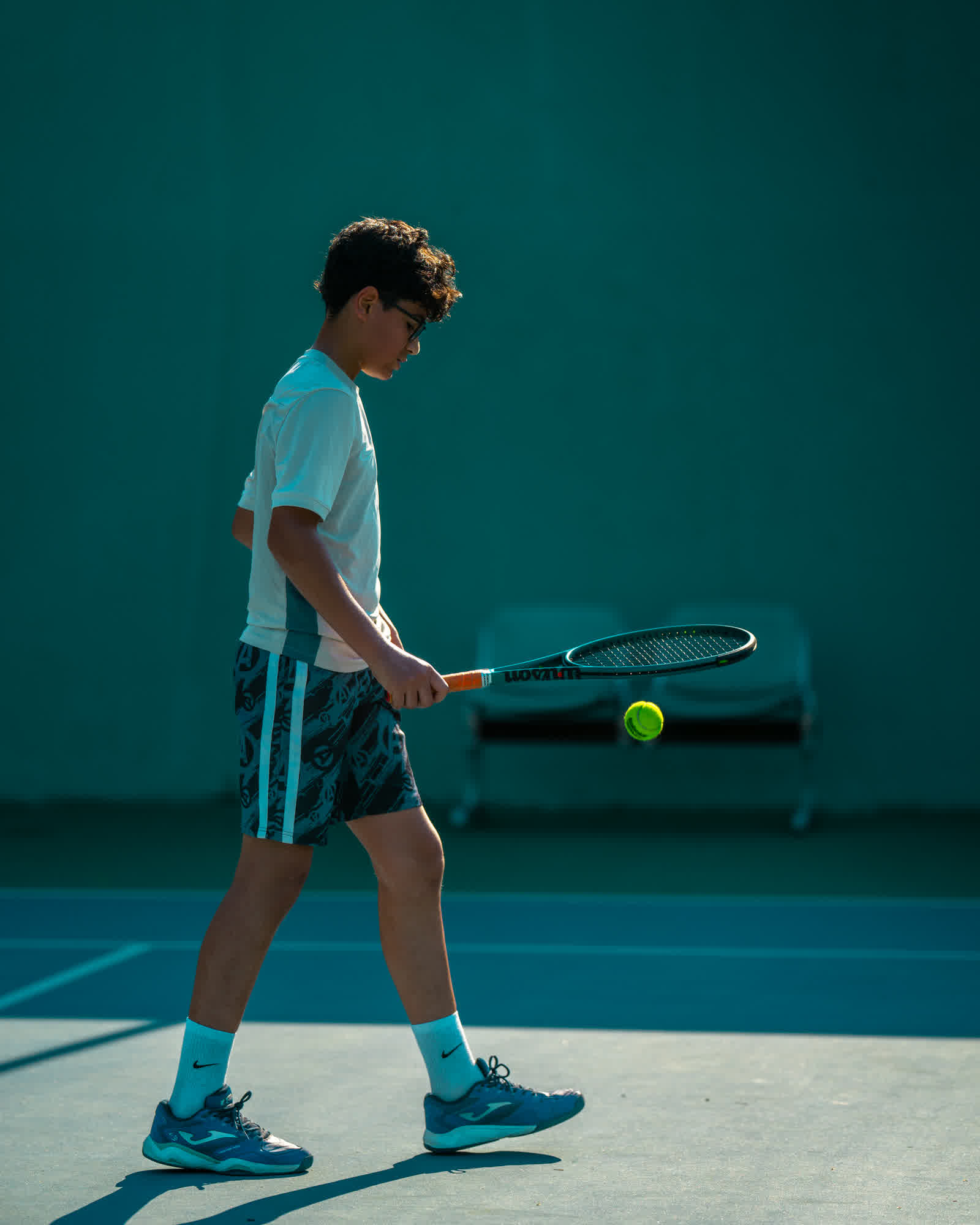 A young player in white standing on a teal court, racket held low.