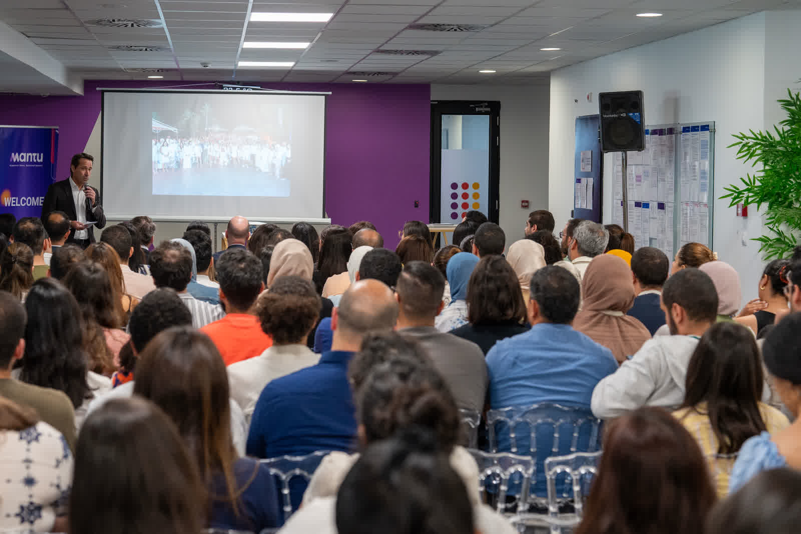 An audience seated in rows facing a presentation screen in a bright conference room.