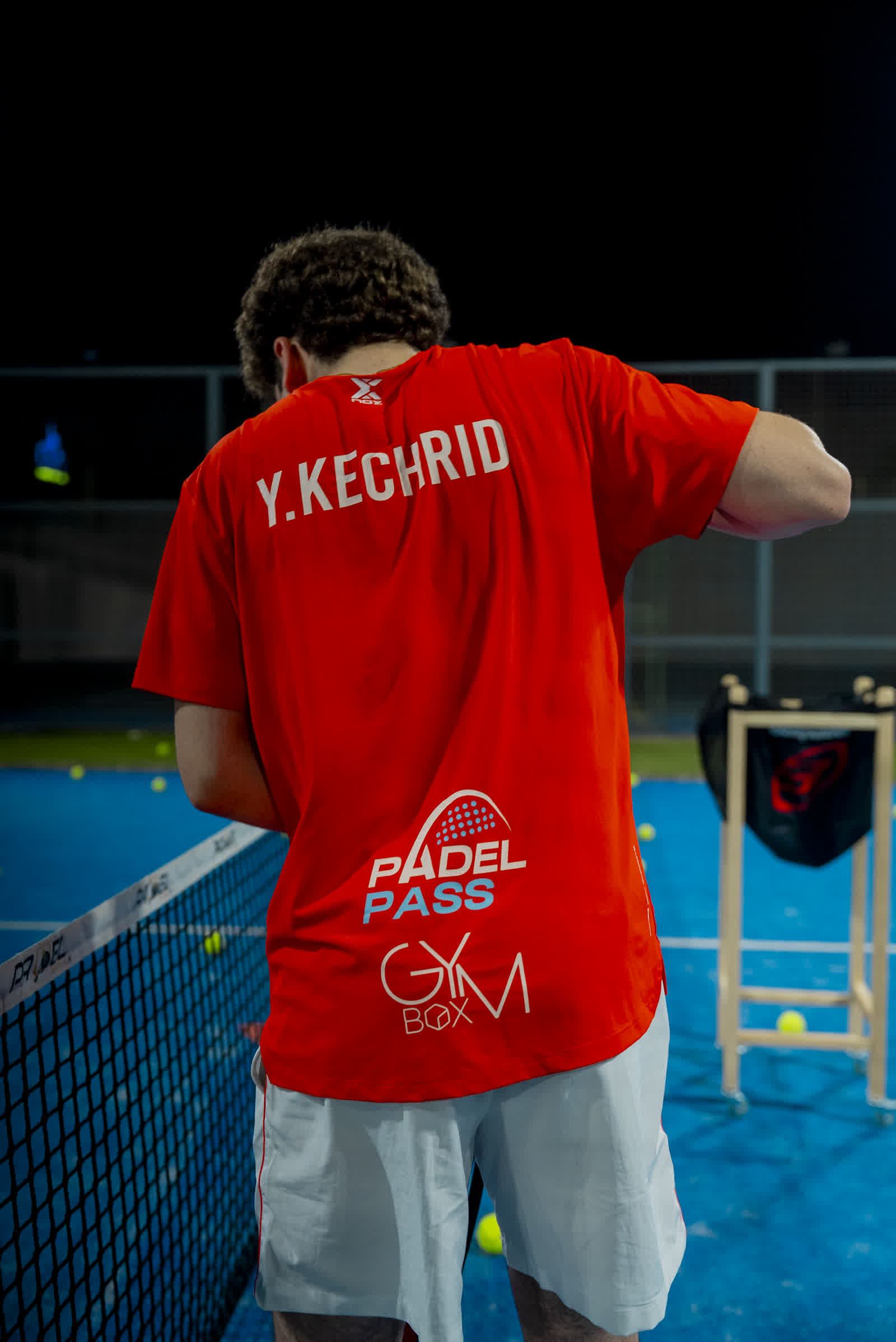 A player in a red shirt at the net on a floodlit court at night, balls scattered at his feet.