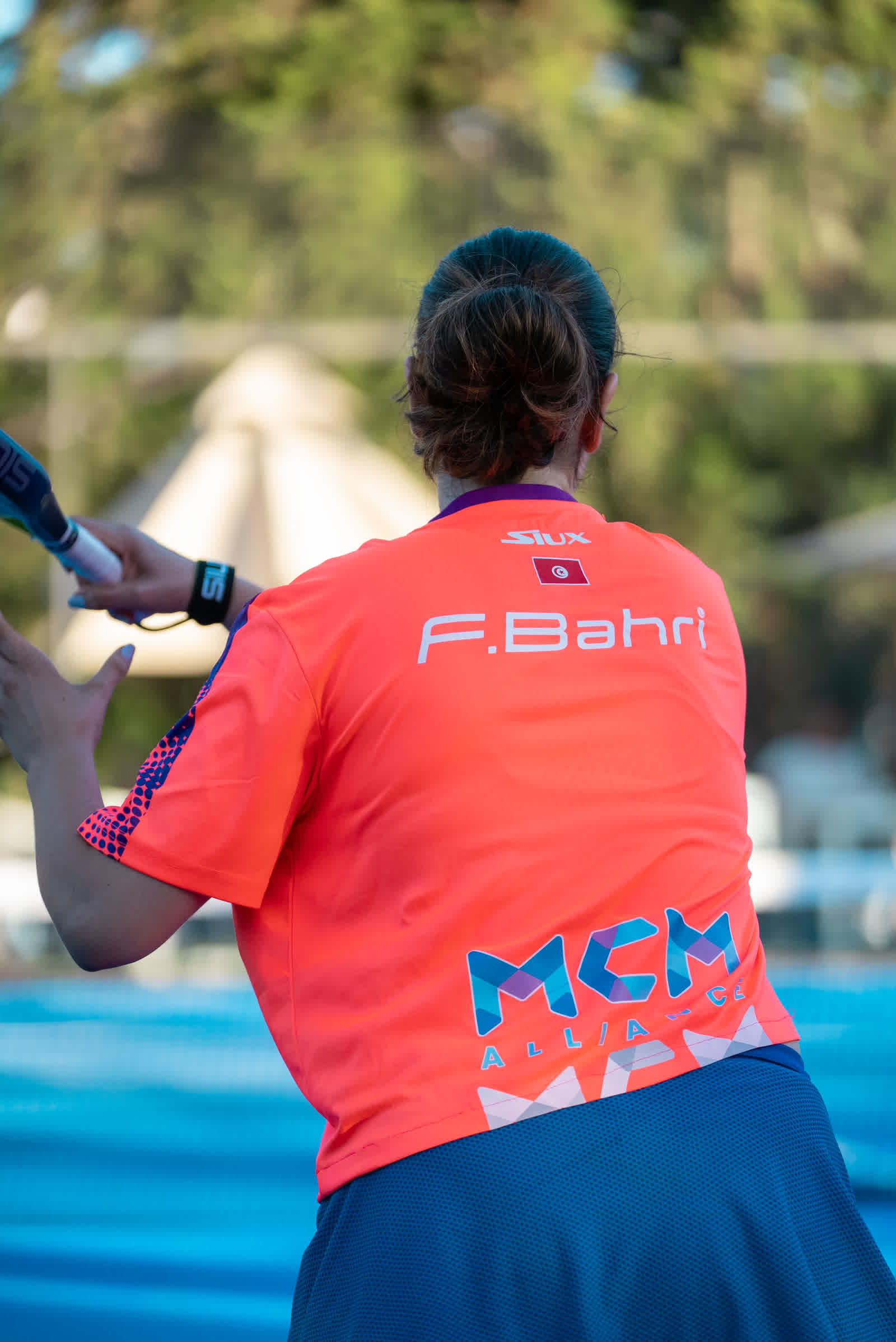 A player in a coral shirt seen from behind, facing the far end of a blue court.