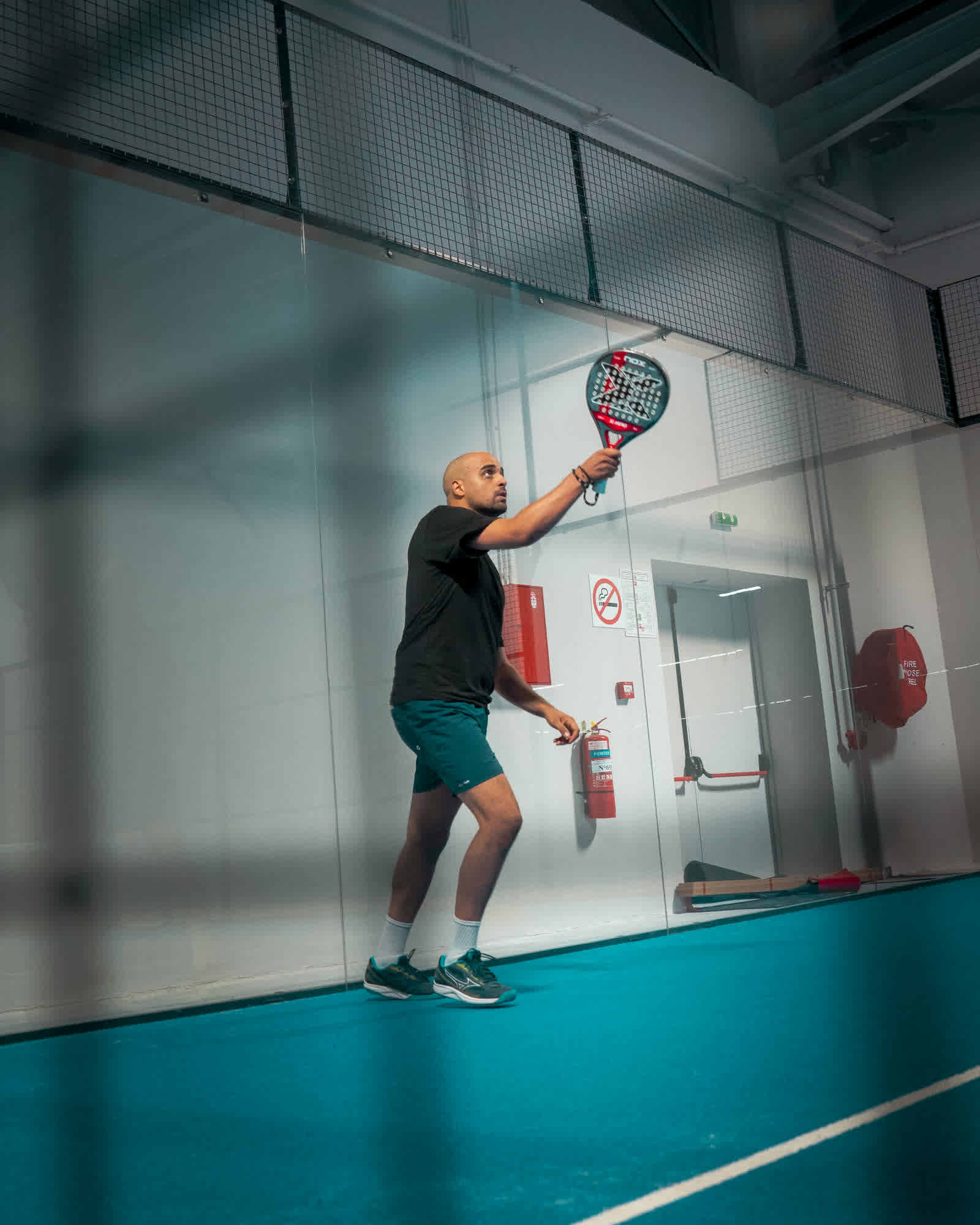 A player reaching up to serve on an indoor court, seen from across the glass.