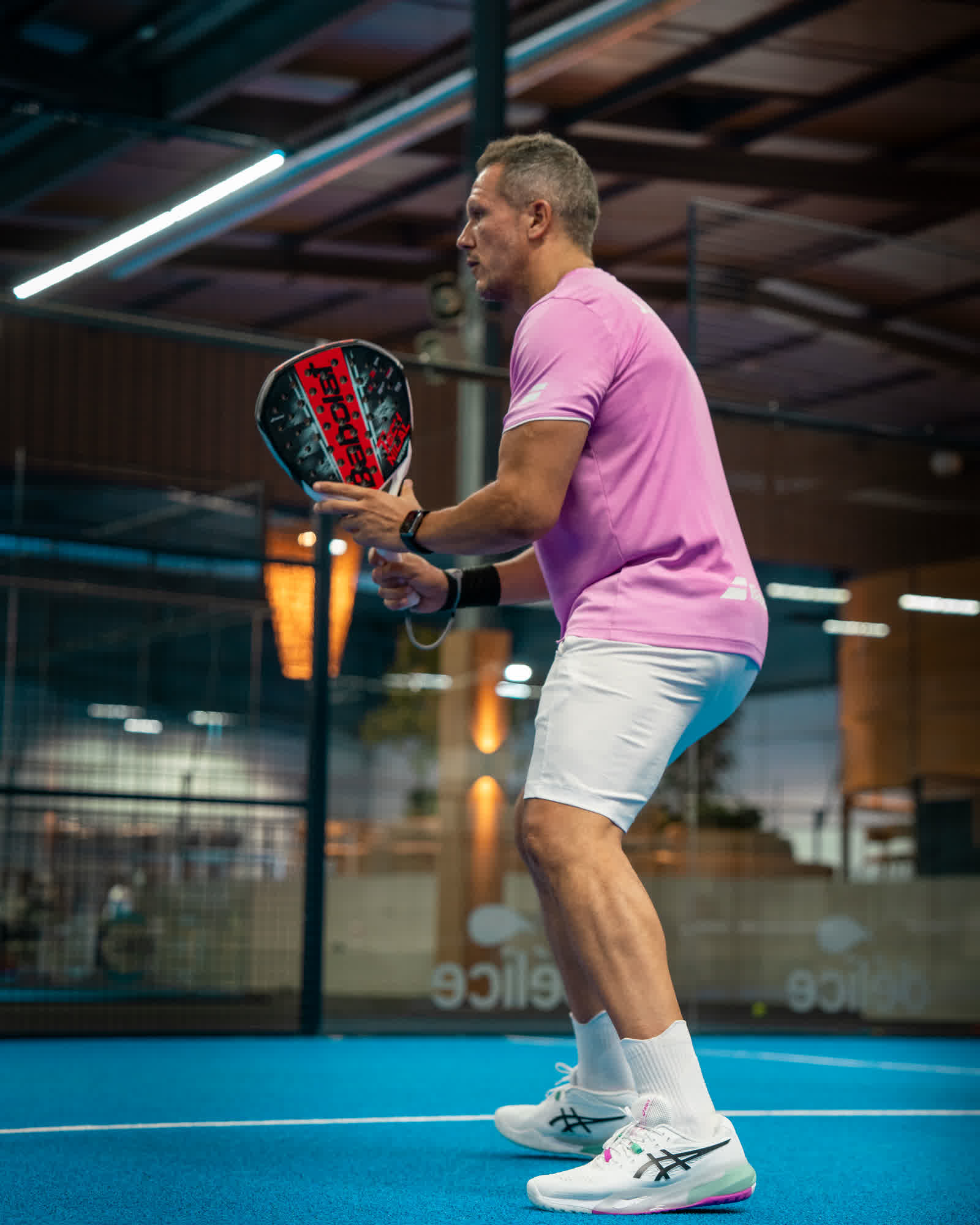 A player in a lilac shirt waiting in a ready stance, racket held in front of him.