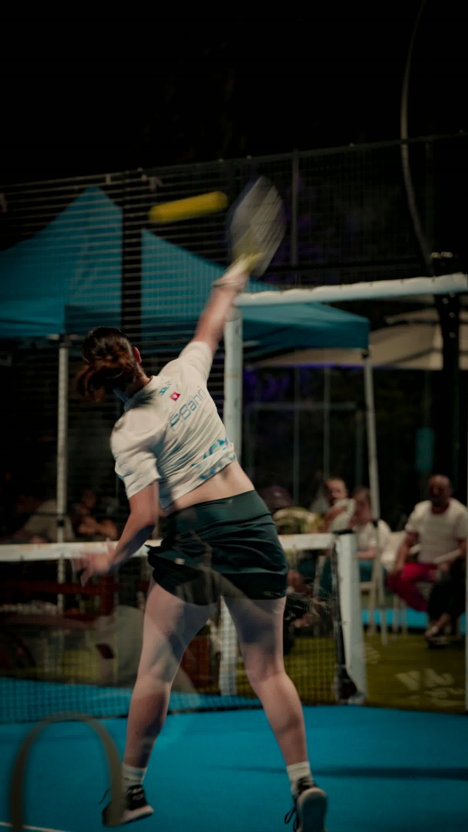 A player stretching up into a smash, arm fully extended above the court lights.
