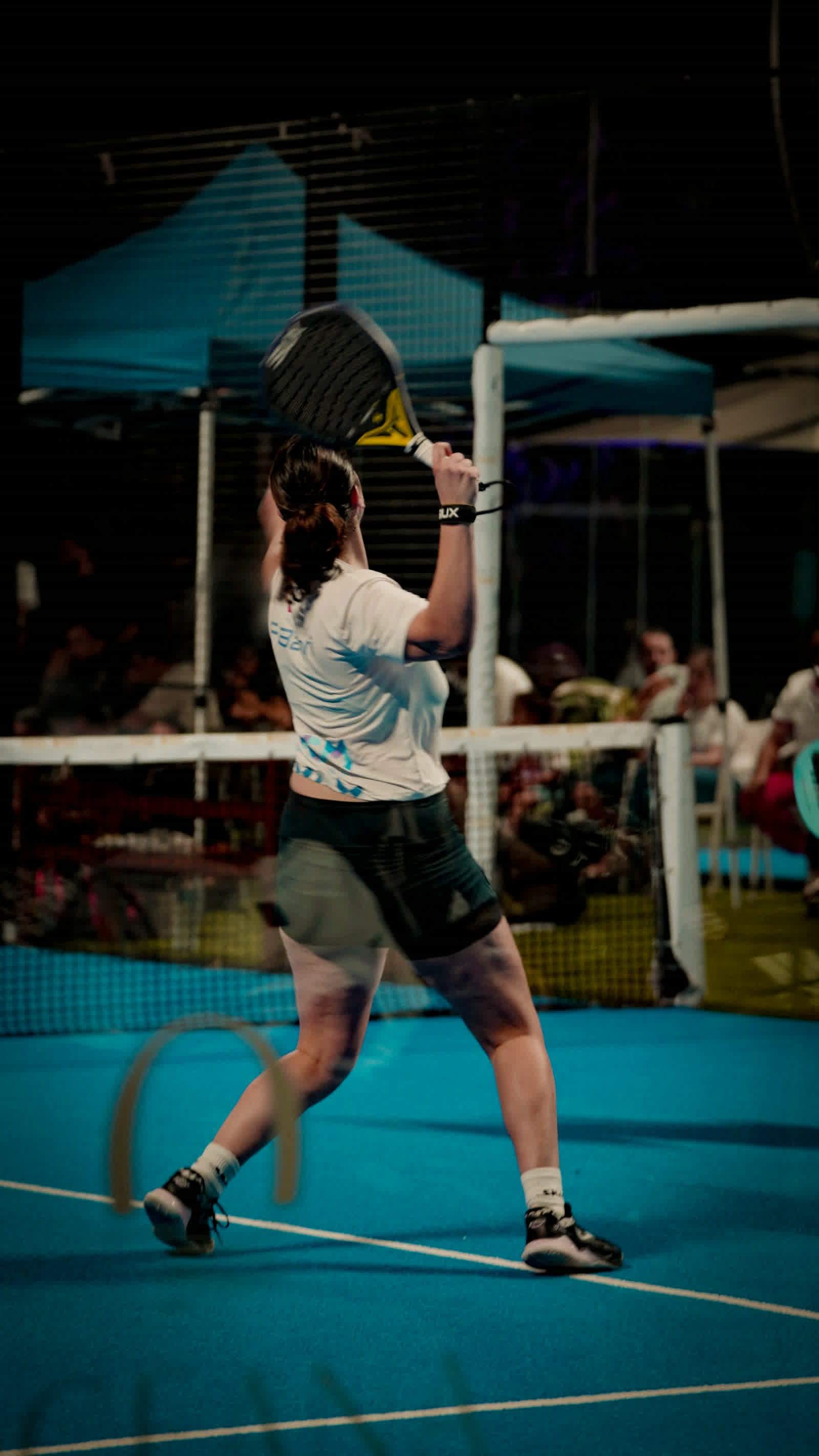 A player driving through a backhand at night, spectators watching from behind the glass.