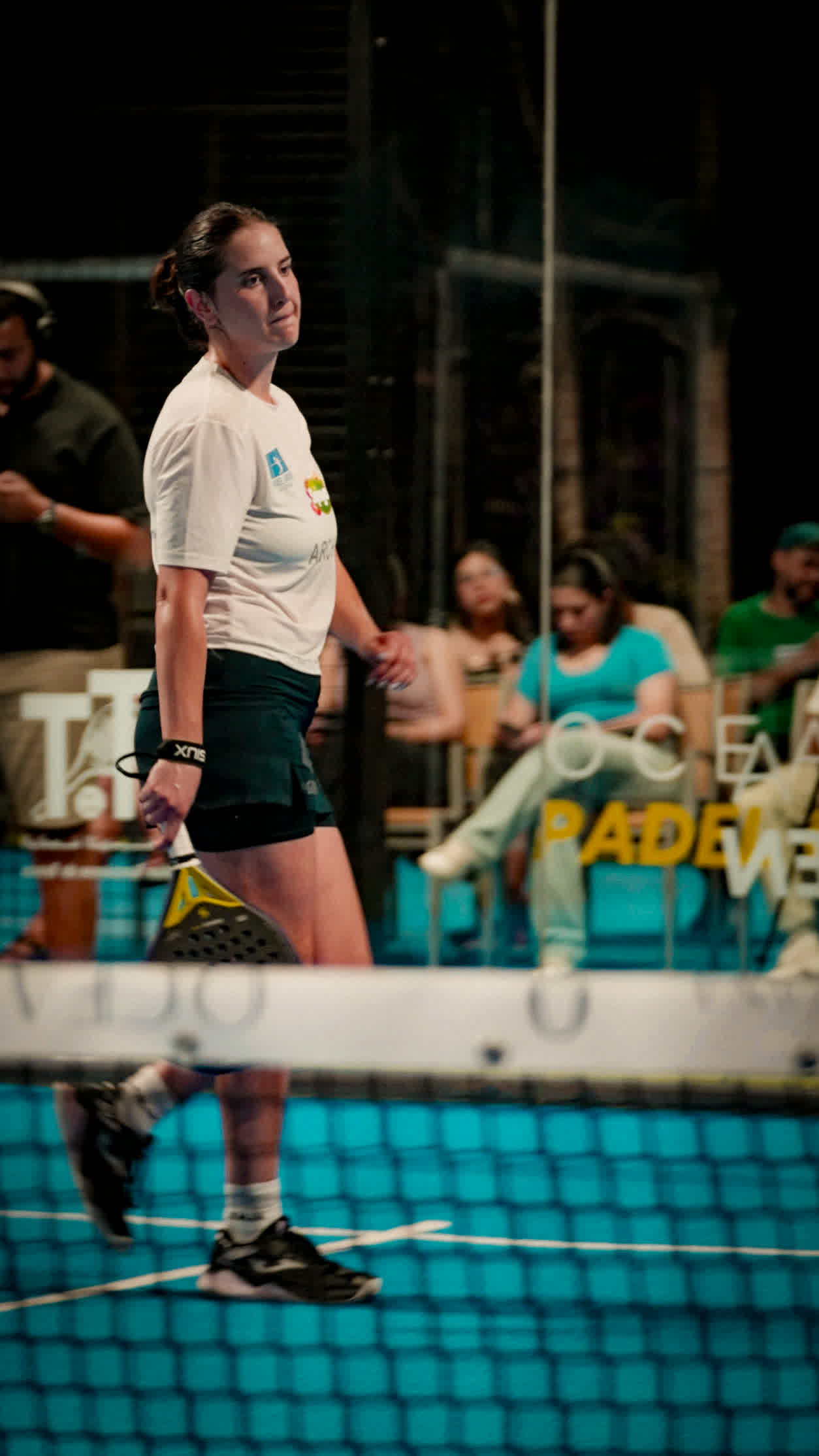 A player at the net under floodlights, a row of spectators seated behind the court.