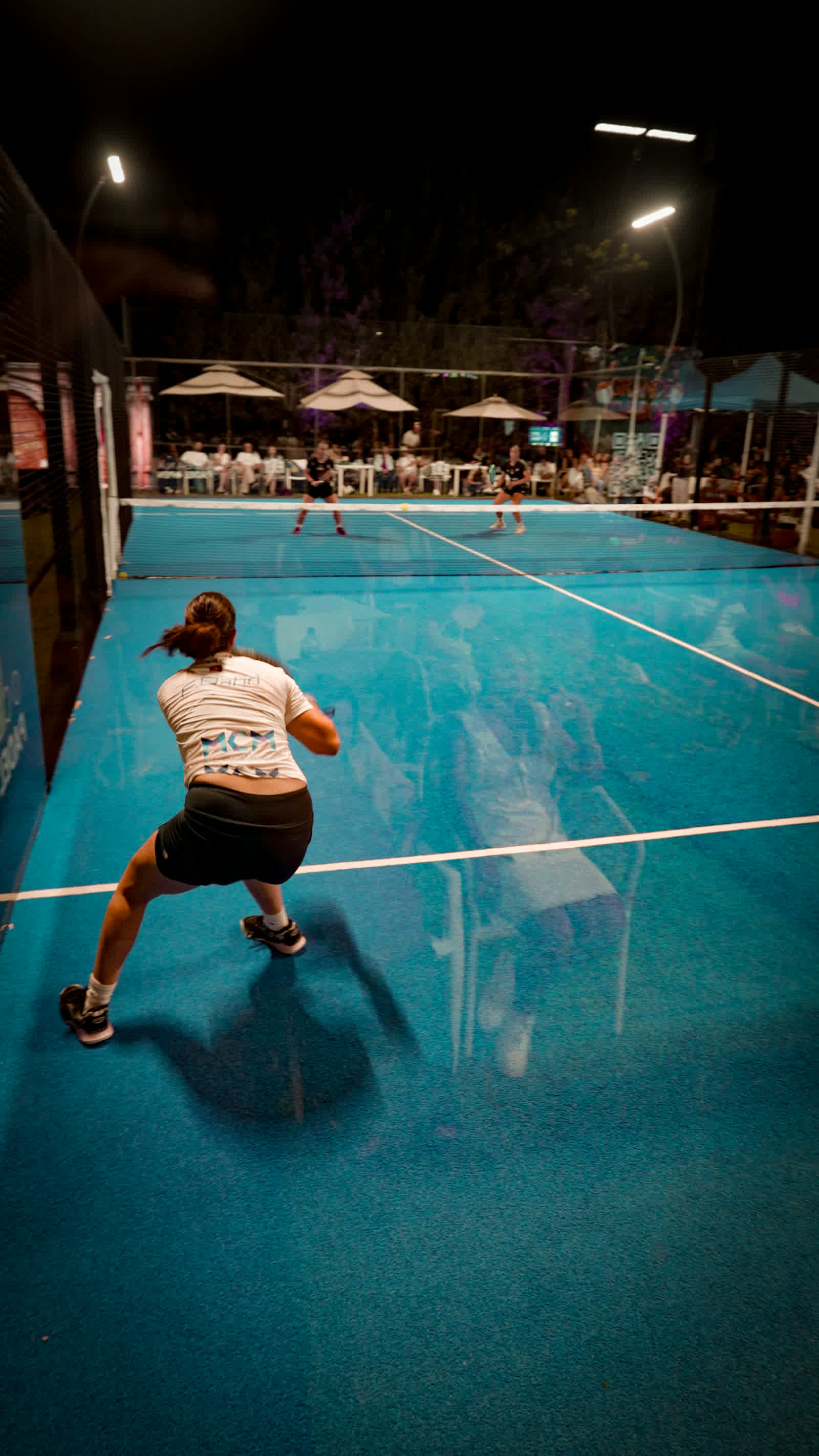 A wide view of a floodlit court at night, a player stretching for a ball near the back glass.